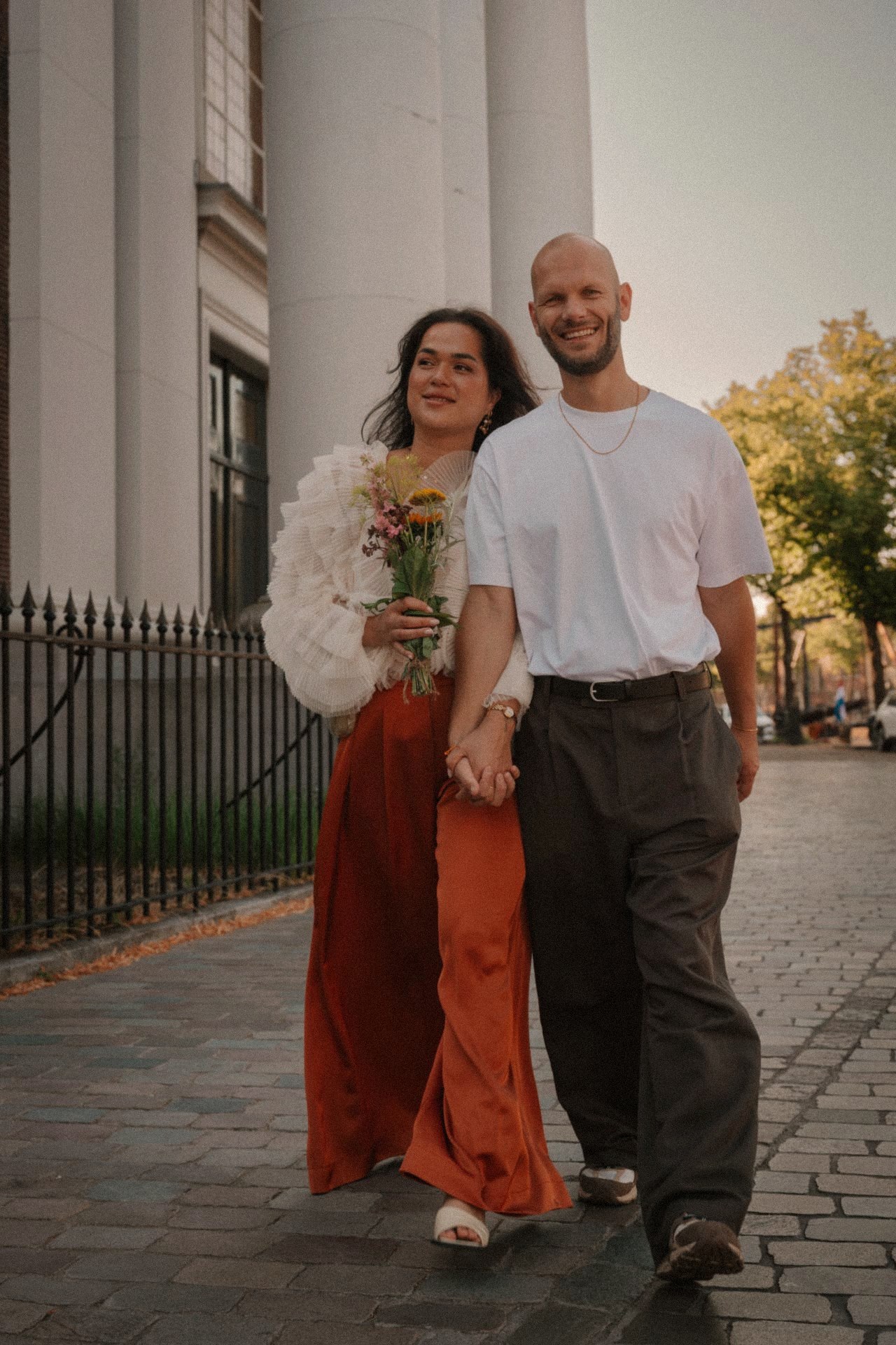 Bruidspaar loopt hand in hand lachend over een keienstraat langs een statig pand